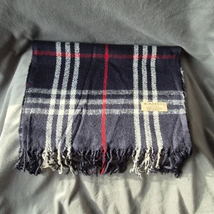 Rutig mörkblå halsduk från Burberry - Snygg halsduk från Burberry i klassiskt rutigt mönster med mörkblå, vita och röda toner. Gjord i 100% lammull och har fransar i kanterna. Perfekt för att ge din outfit en lyxig touch och hålla dig varm under kyliga dagar.