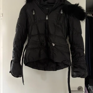 Svart pufferjacka från Rock'n Blue - Svart pufferjacka från Rock'n Blue med fluffig  päls på huvan, flera dragkedjor framtill och justerbart midjebälte. Jackan har quiltad design, långa ärmar och en cool, lite oversized look. Perfekt för kalla dagar när du vill vara både varm och snygg. Använt skick. Pris kan diskuteras 
