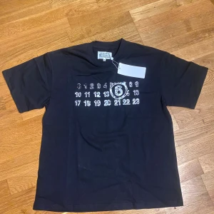 MM6 svart t-shirt - Svart t-shirt från MM6 med ikoniskt siffror-print på bröstet där nummer 6 är markerat. Klassisk rund hals och korta ärmar. Materialet är mjuk bomull och passformen är normal. Perfekt för dig som gillar minimalistisk och edgy design.