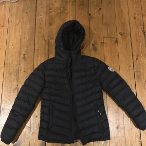 Svart pufferjacka med huva - Svart pufferjacka från Vinston Polo Club  med huva och dragkedja framtill. Jackan har ett quiltat mönster och två sidofickor med dragkedja. Perfekt för kalla dagar och har en stilren look med diskret logga på ärmen.