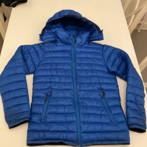 Blå pufferjacka från McKinley 158/164 - Säljer en blå pufferjacka från McKinley i storlek 158/164. Jackan har huva, två dragkedjeförsedda fickor och quiltad design. Yttertyg och foder är i nylon, fyllningen är polyester. Perfekt för kyliga dagar och riktigt skön att bära.
