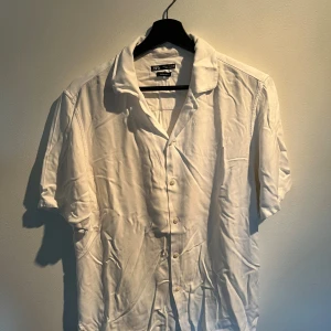 Vit kortärmad skjorta från Zara - Fräsch vit skjorta från Zara med klassisk krage och knappar framtill. Skjortan är kortärmad och har en avslappnad passform, perfekt för varma dagar. Tillverkad i mjuk bomull som känns skön mot huden. Enkel och stilren design som funkar till många olika looks.