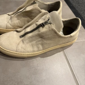 Beige sneakers Axel Arigato mocka - Snygga beige sneakers från Axel Arigato i mjuk mocka med dragkedja framtill istället för klassisk snörning. Skorna har rund tå, platt sula i ljusgult och stilren design. Perfekta för dig som gillar minimalistisk och modern stil.