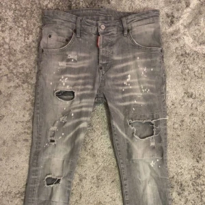 Grå slitna jeans från Dsquared2 - Säljer ett par grå jeans från Dsquared2 med coolt slitna detaljer, stora hål och vit färgstänk över hela byxorna. Modellen är smal och har klassisk femficksdesign. Perfekt för dig som gillar streetstyle och edgy look.
