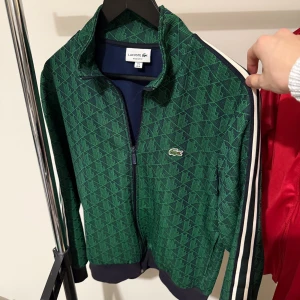 Grön mönstrad vindjacka från Lacoste - Snygg grön vindjacka från Lacoste med geometriskt mönster och klassisk krokodillogga på bröstet. Jackan har vita och svarta ränder längs ärmarna, hel dragkedja och ribbade muddar. Perfekt för dig som gillar sportig och trendig stil.