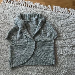 Grå boalero  - Mysig grå stickad väst från Okay med grov kabelstickning, bred krage och en stor knapp framtill. Perfekt att slänga över en långärmad tröja för extra värme. Kortare modell med ribbade kanter och snygg struktur. Storlek 36 passar mig som är xs-s