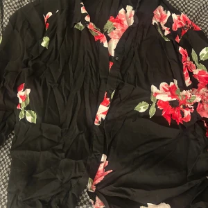 Svart blommig skjorta från H&M - Svart skjorta från H&M med stora blommiga mönster i rött, rosa och vitt samt gröna blad. Skjortan har knappar framtill och lång ärm. Materialet är mjukt och lätt, perfekt för dig som gillar färgstarka prints och vill sticka ut lite extra.