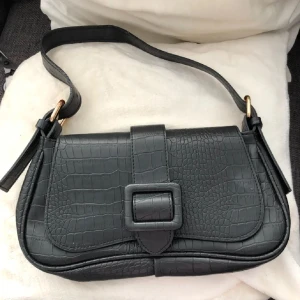 Svart axelväska med krokopräglad yta - Snygg svart axelväska med croco-mönstrad yta och bred axelrem. Väskan har ett spänne framtill och stängs med magnetlås. Insidan är fodrad i svart tyg 