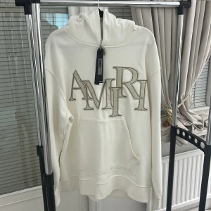 Vit hoodie från Amiri med broderi - Säljer en clean vit hoodie från Amiri med stort broderat logotyptryck på bröstet och en liten broderad detalj på ärmen. Tröjan har huva, känguruficka och långa ärmar. Materialet är mjuk bomull och passformen är normal. Perfekt för dig som gillar streetwear och snygga detaljer.