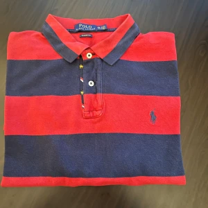 Randig pikétröja Polo Ralph Lauren XXL - Snygg pikétröja från Polo Ralph Lauren i klassisk modell med breda röda och mörkblå ränder. Tröjan har krage, knappar framtill och broderad logga på bröstet. Tillverkad i bomull och passar perfekt för en avslappnad men stilren look.