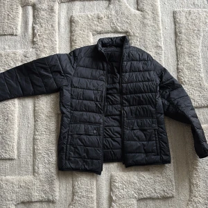 Svart pufferjacka från Barbour - Svart pufferjacka från Barbour med raka quiltade paneler, hög krage och två stora fickor med tryckknappar framåt. Den har bra passform och passar dig som är runt 160-180