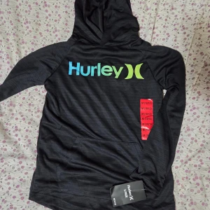Svart hoodie från Hurley med logga - Svart hoodie från Hurley med stor färgglad logga i blått och grönt på bröstet. Tröjan har huva, lång ärm och en klassisk känguruficka framtill. Perfekt för en chill och sportig stil.