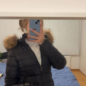 Svart pufferjacka med päls från Rockandblue - Svart pufferjacka från Rockandblue med fluffig brun fuskpäls på huvan. Jackan har dragkedja framtill, quiltad design och långärmade ärmar. 