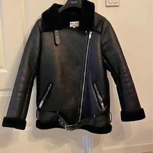 Svart pälsjacka från NA-KD - Säljer en svart pälsjacka från NA-KD med bred krage och mjuk fuskpäls på insidan. Jackan har två sneda fickor med dragkedja, bälte nedtill och detaljer i silver. Perfekt för kalla dagar och har en cool, oversized vibe.