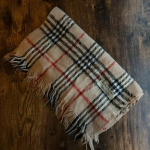 Burberry Halsduk - Elegant Burberry-halsduk i 100% lammull, tillverkad i England. Klassisk design med det välkända rutmönstret. Mycket bra skick, små fläckar eller utan hål. Ett tidlöst plagg som lyfter varje outfit.