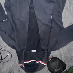 Mörkblå Moncler hoodie med dragkedja - Snygg mörkblå hoodie från Moncler med hel dragkedja och huva. Tröjan har Moncler-logga på bröstet och coola ribbade muddar med röd, vit och blå rand. Tillverkad i mjuk bomull, perfekt för chill dagar. Två fickor framtill och stilren design.