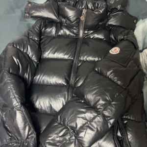 Svart Moncler pufferjacka med huva - Svart pufferjacka från Moncler med glansig finish och stor huva. Jackan har Moncler-logga på ärmen, dragkedja framtill och breda, quiltade sektioner. Perfekt för kalla dagar och har en riktigt snygg streetstil. Har ett håll har inget kvitto då jag köpte för länge sen därför säljer jag billigt