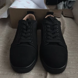 Svarta sneakers från Christian Louboutin - Stilrena svarta sneakers från Christian Louboutin med klassisk röd sula. Skorna har rund tå, snörning och är tillverkade i skinn med en matt finish. Insidan är ljusbeige och sulan har den ikoniska Louboutin-signaturen. Perfekta för dig som gillar exklusiv streetstyle.