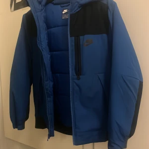 Blå pufferjacka från Nike med huva - Snygg blå pufferjacka från Nike med svarta detaljer och huva. Jackan har dragkedja framtill, en bröstficka med dragkedja och vadderad insida för extra värme. Perfekt för kalla dagar och har en sportig look med Nike-logga på bröstet.
