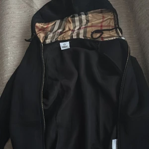 Svart vindjacka med mönstrat foder - Svart vindjacka från Burberry med klassiskt randigt innerfoder i beige, rött och grått. Jackan har huva med dragsko och dragkedja framtill. Perfekt för dig som vill ha en stilren jacka med lyxig känsla och snygga detaljer.