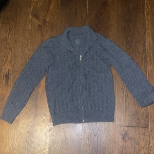 Grå stickad kofta från D&G Junior - Mysig grå stickad kofta från D&G Junior med kabelstickat mönster och hel dragkedja framtill. Hög krage och diskret logga i nacken. Perfekt för lager-på-lager under kyliga dagar.