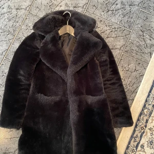 Svart pälsjacka från Amisu - Mysig svart fuskpälsjacka från Amisu med bred krage och två stora fickor framtill. Jackan har en klassisk rak passform och är fodrad på insidan för extra värme. Perfekt för kalla dagar när du vill ha en stilren look.