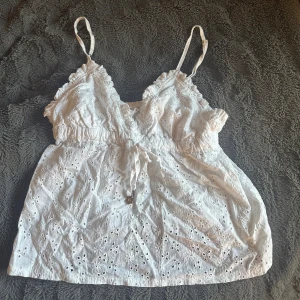 Vit broderad linnetopp  - Supersöt vit linnetopp från Censored, New Yorker i storlek XXS. Toppen har broderade blommor, justerbara smala axelband och en liten rosett framtill. Den har v-ringning, volangdetaljer och resor i midjan. Perfekt för varma dagar och ger en romantisk vibe.