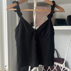 Svart linnetopp med snörning Divided - Säljer en svart linnetopp från Divided med breda axelband och snygg snörning både fram och bak. Toppen har en lätt v-ringning och är tillverkad i ett mjukt polyestertyg. Perfekt för dig som gillar en enkel men ändå trendig look.