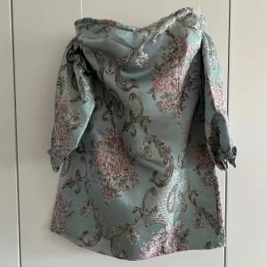 Blommig kortklänning med puffärm - Superfin kortklänning i ljusblått med blommigt mönster i rosa och beige toner. Klänningen har puffiga trekvartsärmar och offshoulder-skärning. Materialet känns glansigt och lyxigt, perfekt för dig som vill sticka ut med en romantisk vibe.