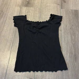 Svart ribbad topp från Hollister XS - Svart ribbad topp från Hollister med söt rynkdetalj framtill och volangkant vid halsringning, ärmslut och nederkant. Modellen är figurnära och har korta ärmar med offshoulder-stil. Perfekt för dig som gillar en enkel men ändå trendig look.