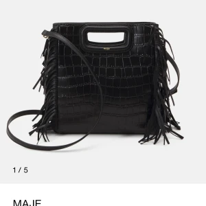 Svart axelväska med fransar från Maje - Stilren svart axelväska från Maje i croco-präglat skinn med coola fransdetaljer på sidorna. Väskan har både handtag och lång axelrem, perfekt för att bära på olika sätt. Snygg och edgy design som lyfter vilken outfit som helst.