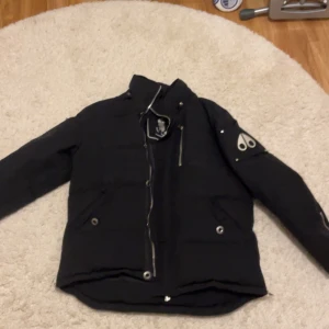 Svart pufferjacka från Moose Knuckles - Säljer en svart pufferjacka från Moose Knuckles med ikonisk logga på ärmen. Jackan har dragkedja, knappar och två fickor framtill. Fodrad och perfekt för kalla dagar. Snygg och stilren design som passar till det mesta.