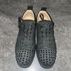 Svarta sneakers med nitar från Louboutin - Säljer ett par svarta sneakers från Christian Louboutin med mattsvarta nitar på tån och klassisk röd sula. Skorna är i mocka med svarta snören och har en riktigt edgy vibe. Insidan är beige och de har rund tå. Perfekta för dig som vill sticka ut.