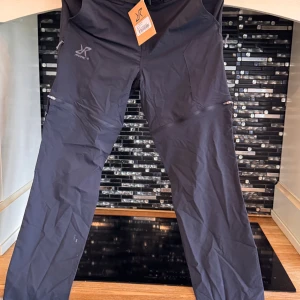 Svarta täckbyxor från RevolutionRace - Svarta täckbyxor från RevolutionRace med raka ben, praktiska fickor med dragkedja och justerbart bälte i midjan. Tillverkade i slitstarkt syntetmaterial, perfekta för utomhusaktiviteter. Snygg och funktionell design med diskret logga på låret.