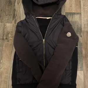 Mörkblå cardigan med huva från Moncler - Mörkblå cardigan från Moncler med huva och dragkedja framtill. Jackan har quiltade detaljer på sidorna, ribbade muddar och Moncler-logga på vänster ärm. Materialmix med polyester på kroppen och bomull på ärmarna ger en schysst kontrast.