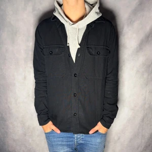 Svart overshirt från Mads Nørgaard - Svart overshirt från Mads Nørgaard med klassisk krage, två bröstfickor med lock och knappar framtill. Skjortan har en rak passform och är tillverkad i bomull, perfekt för lager-på-lager-stil.