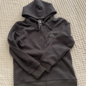 Grå hoodie från Lacoste med dragkedja - Snygg grå hoodie från Lacoste med klassisk krokodillogga på bröstet. Tröjan har huva med snörning, långa ärmar och praktisk dragkedja framtill. Perfekt för chill dagar och enkel att matcha med jeans eller joggers.