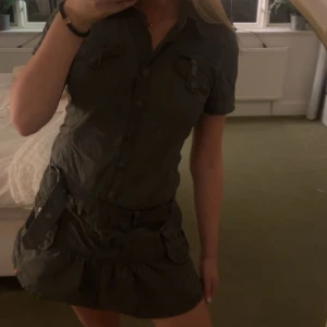 Mörkgrå kort klänning från edc - Snygg mörkgrå kort klänning från edc med skjortkrage, knappar framtill och korta ärmar. Klänningen har fickor på bröstet och kjolen, samt ett matchande bälte i midjan. Perfekt för dig som gillar utility-stil och vill ha något bekvämt men ändå coolt. Priset går att diskutera!