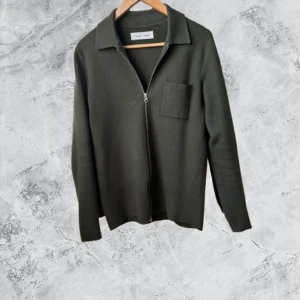 Mörkgrön kofta från Samsøe Samsøe - Stilren mörkgrön kofta från Samsøe Samsøe med dragkedja framtill, klassisk krage och en bröstficka. Koftan är långärmad och tillverkad i ett mjukt material som passar perfekt till lager-på-lager. Enkel och clean design för en snygg look.