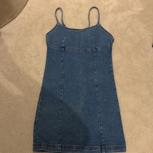 Blå jeansklänning med smala axelband - Säljer en blå jeansklänning med smala axelband och figurnära passform. Klänningen är kort och har en dragkedja i ryggen för enkel påklädning. Snygga sömdetaljer framtill ger en cool och stilren look. Perfekt för dig som gillar denim och vill ha en enkel men trendig klänning.