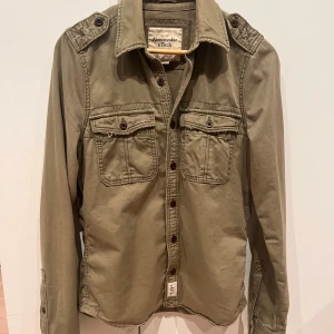 Olivgrön overshirt från Abercrombie & Fitch - Olivgrön overshirt i M (-L) från Abercrombie & Fitch med coola axeldetaljer, två bröstfickor med knappar och tryck 'A&F 92' på ryggen. I använt men gott skick! Hör av er vid funderingar.