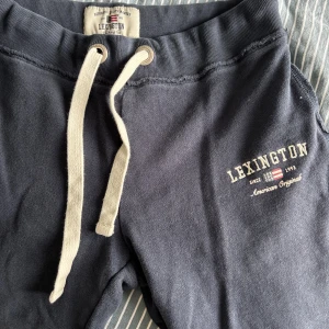 Mörkblå mjukisbyxor från Lexington - Mörkblå mjukisbyxor från Lexington med vit dragsko i midjan och broderad logga på ena benet. Byxorna har raka ben med mudd nertill och är tillverkade i mjuk bomull. Hål på mudden syns på tredje bilden. Passar Xs- S