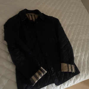 Svart quiltad jacka från Burberry - Snygg svart quiltad jacka från Burberry med klassiskt rutigt foder i beige, rött och svart. Jackan har knäppning framtill, krage och långa ärmar med uppvik där det rutiga fodret syns. Perfekt för dig som vill ha en stilren och tidlös look.