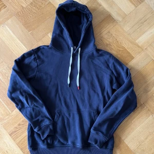 Mörkblå hoodie med huva och ficka från Les Deux - En stilren mörkblå hoodie från Les Deux med huva och vita snören med les deux färgerna på ändarna. Perfekt för dig som gillar en enkel och clean look.