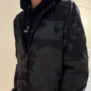 Camouflage jacka från Woolrich - Säljer en snygg jacka från Woolrich i mörk camouflage. Jackan har dragkedja framtill, Perfekt för dig som vill sticka ut med streetstyle-vibe. Färgskalan går i svart och mörkgrått.