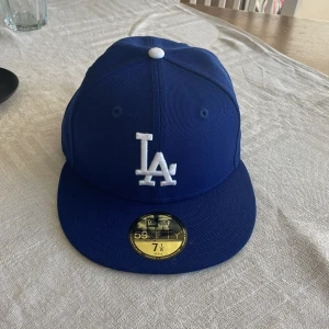 Blå LA Dodgers keps från New Era - Snygg blå 59FIFTY keps från New Era med LA Dodgers-logga broderad i vitt framtill. Klassisk flat peak och sticker på skärmen. Tillverkad i 100% polyester och officiell MLB on-field cap. Perfekt för dig som gillar sportig streetstyle. Helt oanvänd.