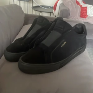 Svarta sneakers från Axel Arigato - Säljer ett par helsvarta sneakers från Axel Arigato i mocka med diskret guldtext på sidan. Skorna har en minimalistisk design utan synlig snörning och svart platt sula. Insidan är fodrad med mjukt skinn för extra komfort. Perfekta för en clean och stilren look. PRIS KAN DISKUTERAS 