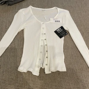 Vit ribbad topp med knappar NLY Trend XS - Vit ribbad topp från NLY Trend i storlek XS. Modellen har långa ärmar, v-ringning och silverfärgade knappar framtill. Tunt och stretchigt material som sitter snyggt på kroppen. 