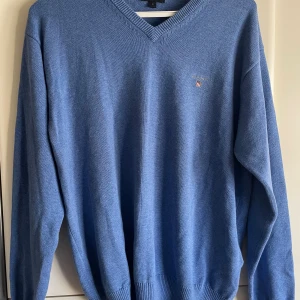 Gant v-ringad tröja blå - Säljer en klassisk blå v-ringad tröja från GANT med broderad logga på bröstet. Tröjan är långärmad och har ribbade muddar vid ärmslut och nederkant. Perfekt för lager-på-lager och enkel att matcha med jeans eller chinos. Stl L