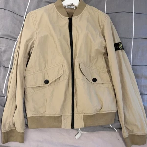 Beige bomberjacka från Stone Island - Säljer en beige bomberjacka från Stone Island med klassisk svart logotyp-patch på ärmen. Jackan har två stora fickor med knappar, ribbade muddar och dragkedja framtill. Materialet är lätt och syntetiskt, perfekt för vår och höst. Stilsäker och clean look.